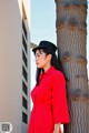 A woman in a red coat and hat leaning against a palm tree.